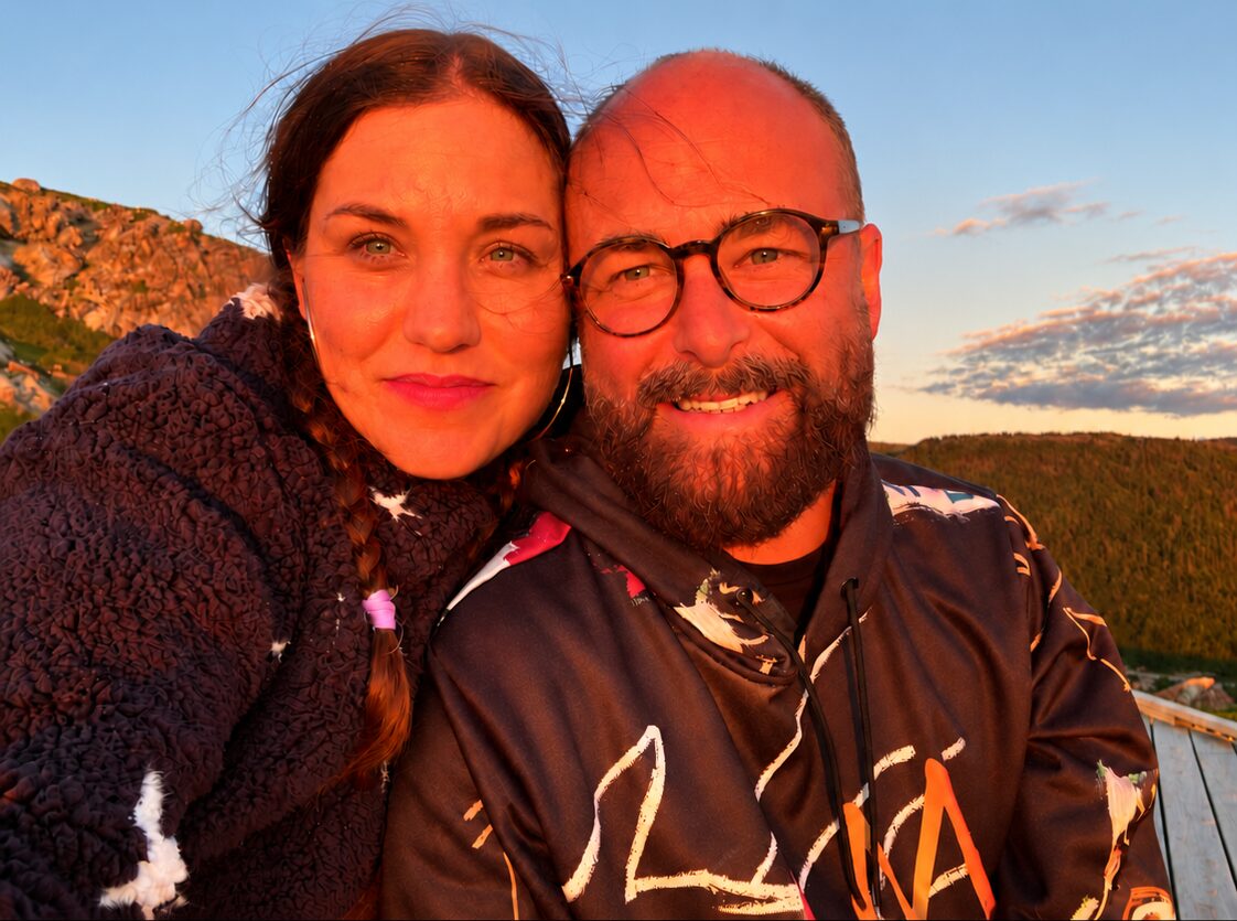 Nina and John cheek to cheek at sunset in Nova Scotia, on the trip where Cliick started, with a hillside and treeline behind them